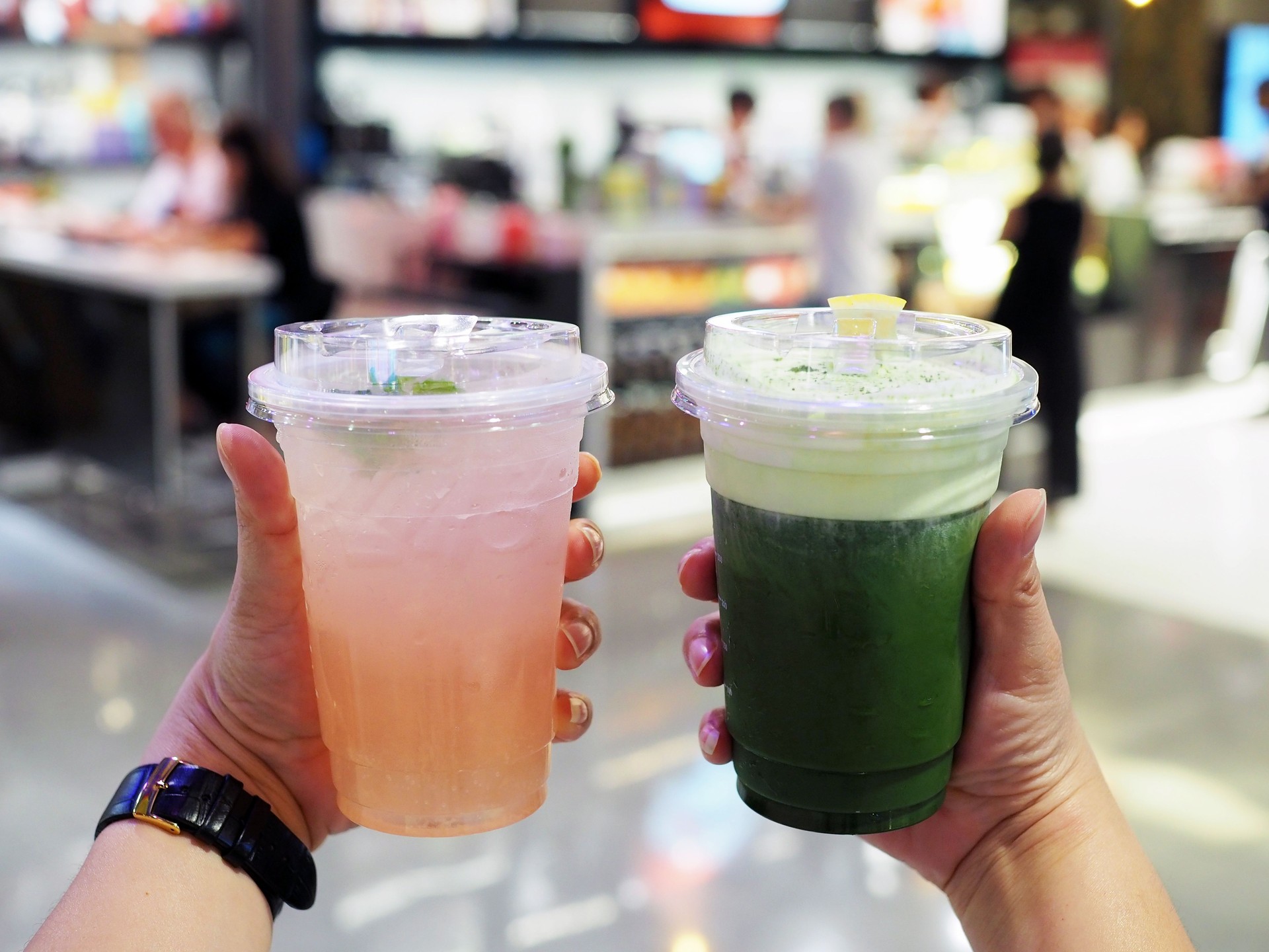Hands holds a plastic glass of iced peach soda with crystal bubble and iced green tea latte with layer of fresh foam milk on blurred bokeh background.