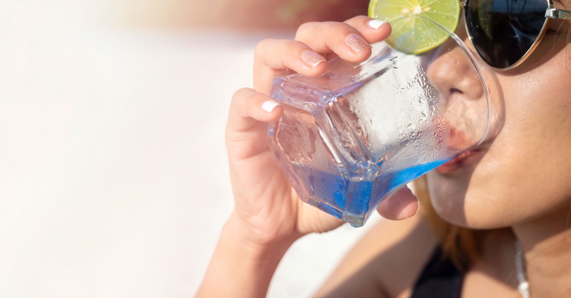 Close-up woman's hand with glitter fashionable nail polish holding blue cocktail drink with lime in the short glass, welcome drink with copy space. Summer banner background. Woman with sunglasses.